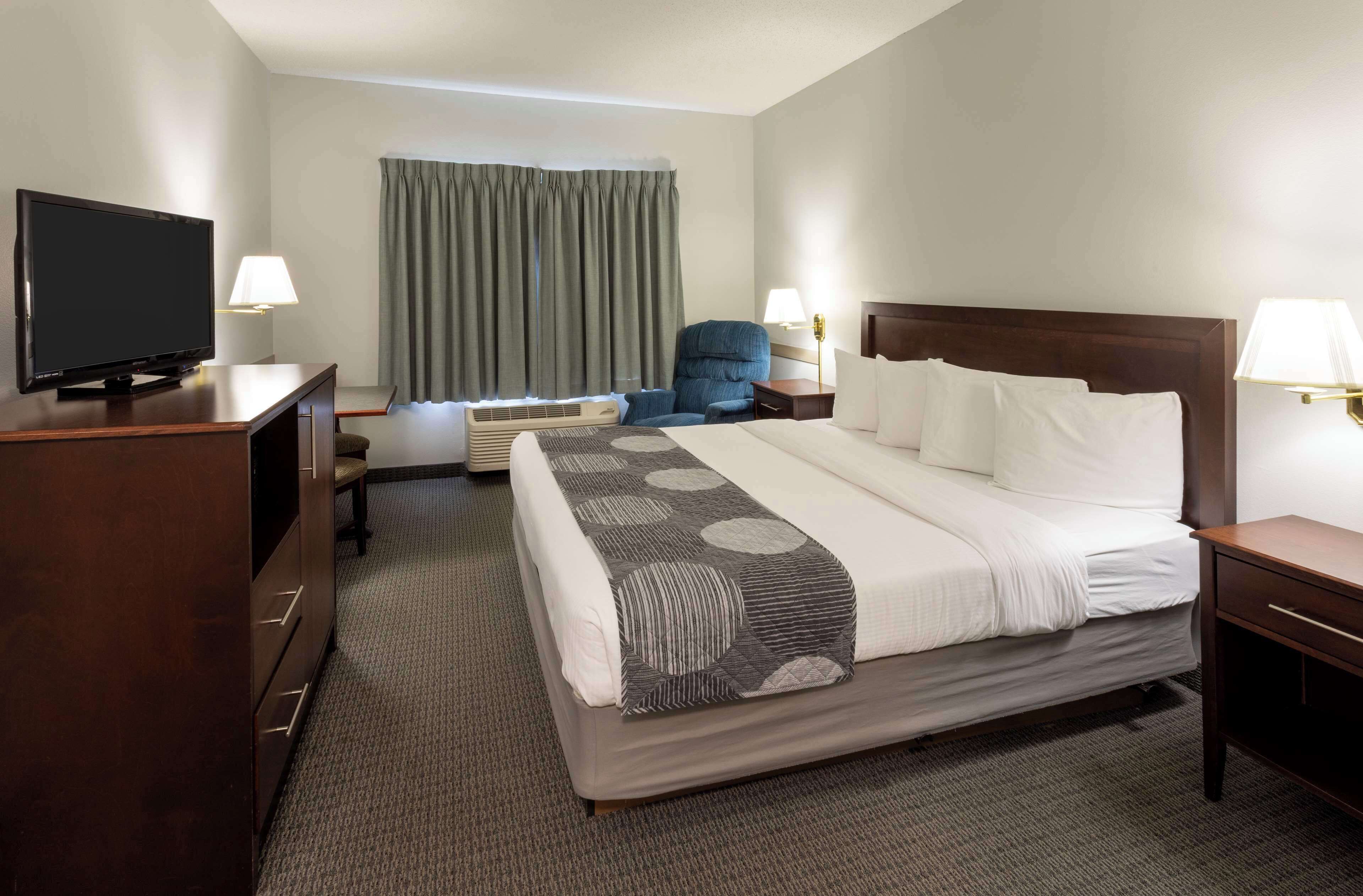 A hotel room with a bed, a desk with a television, a chair, a lamp, and a window with curtains.