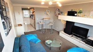 A modern living room with a sofa, coffee table, and TV stand. There is a kitchen in the background with a window and a decorative wreath. - Parklife Apartments (Winterberg)