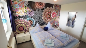A bedroom with a colorful tapestry on the wall, a bed with a checkered duvet, and a bedside table with a lamp. - Parklife Apartments (Winterberg)