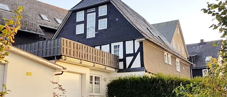 Uma casa tradicional em estilo de enxaimel com telhado inclinado, varanda de madeira e um hedge bem cuidado na frente.