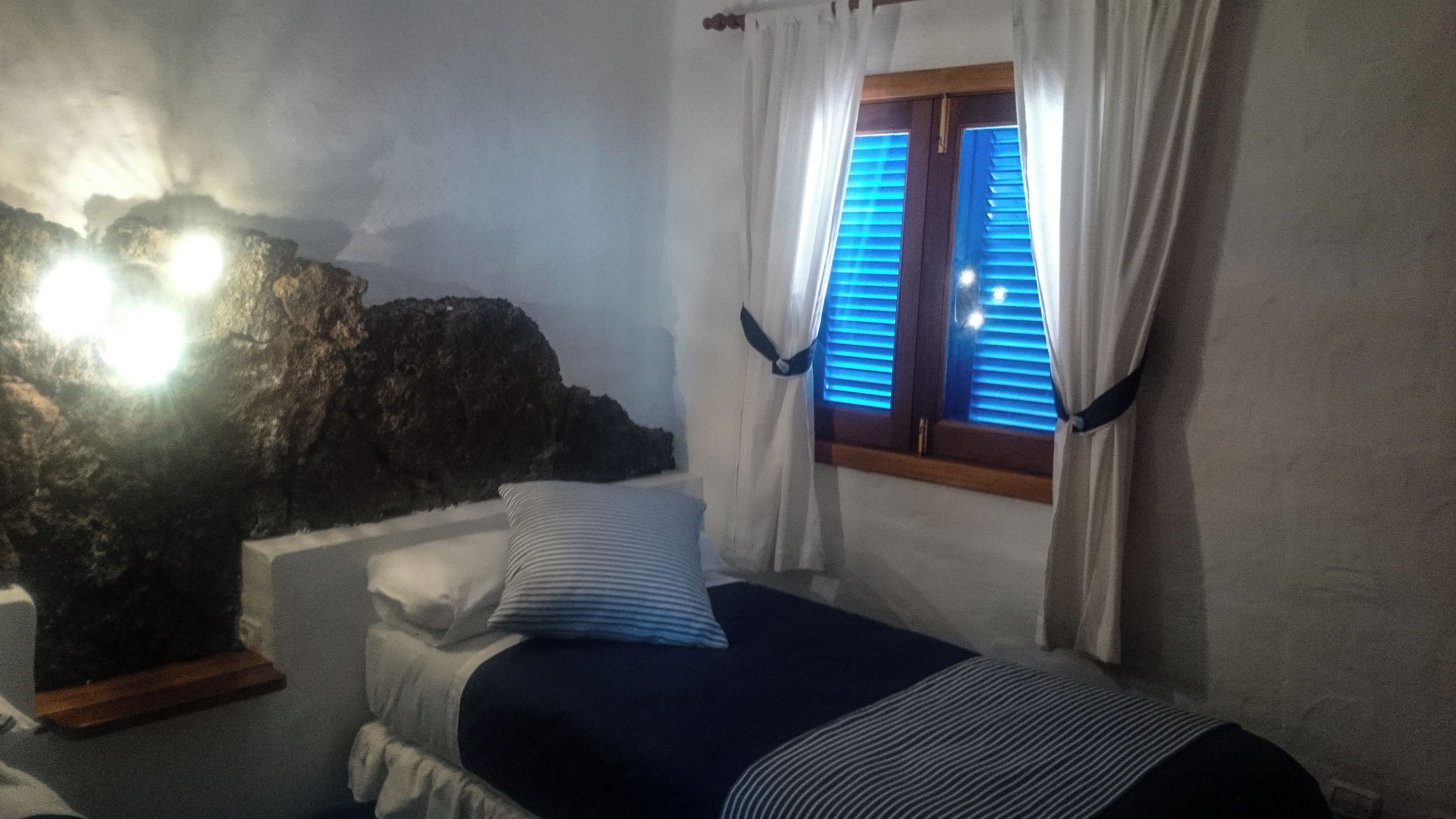 Una habitación con cama, ventana con contraventanas azules y pared con textura similar a roca.