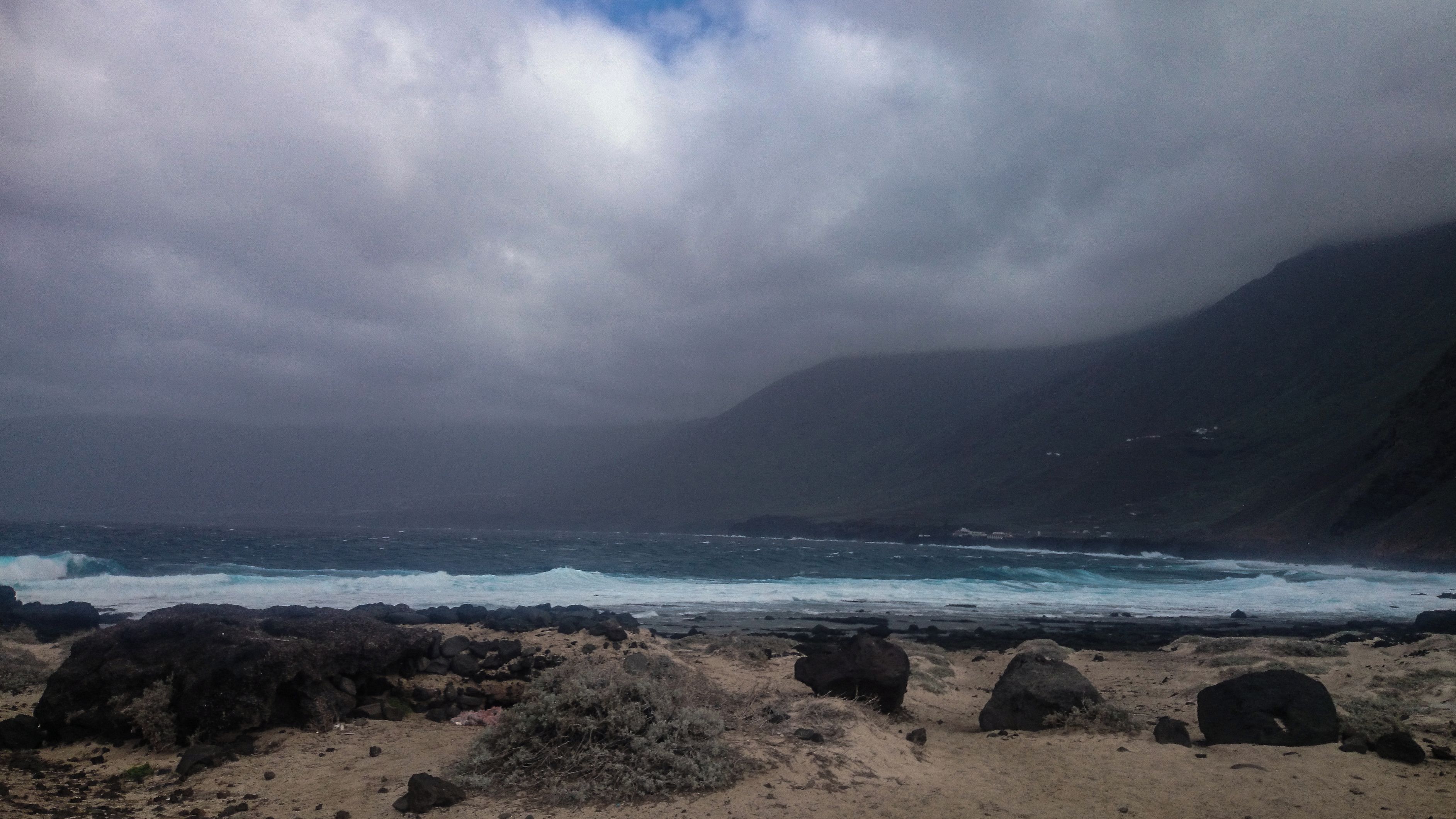 Un paisaje costero con playa rocosa, olas y una montaña al fondo.