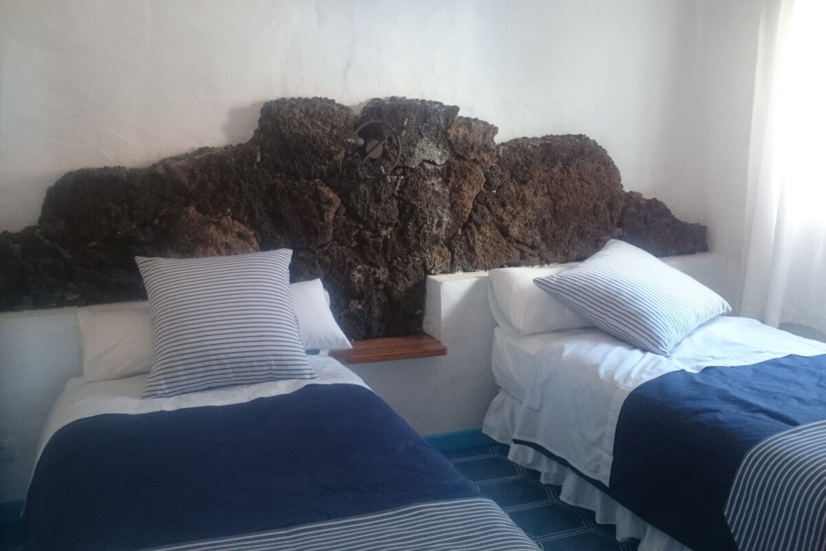 A room with two beds, each with a striped pillow and a blue blanket. The walls are plain with a textured rock-like feature above the beds.