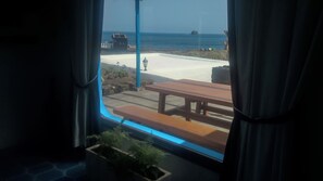 A view from a window with a wooden bench and a potted plant, overlooking a sandy beach and a body of water with a building in the distance. - Villa Juan (Frontera)