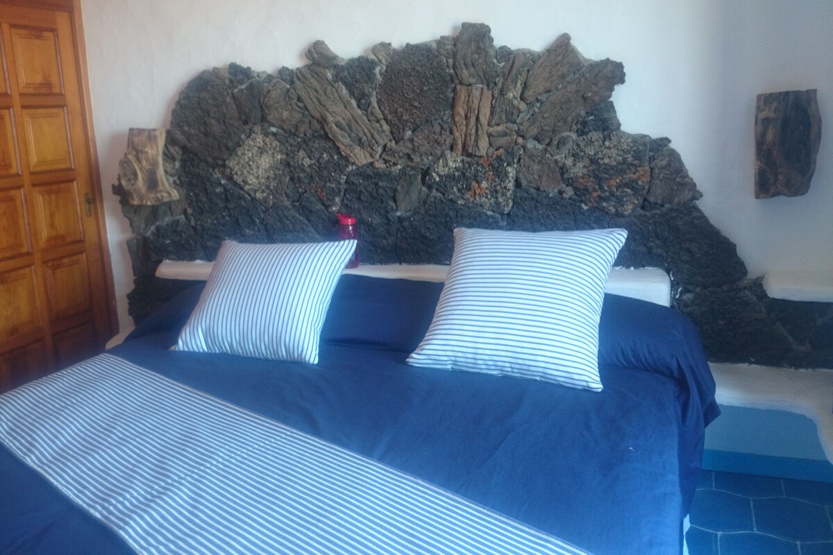 Una cama con colcha azul y blanca a rayas, dos almohadas y cabecero de piedra.