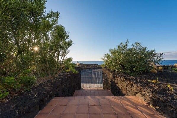 Un sendero conduce a una playa con una reja de metal, rodeado de vegetación y un cielo despejado.