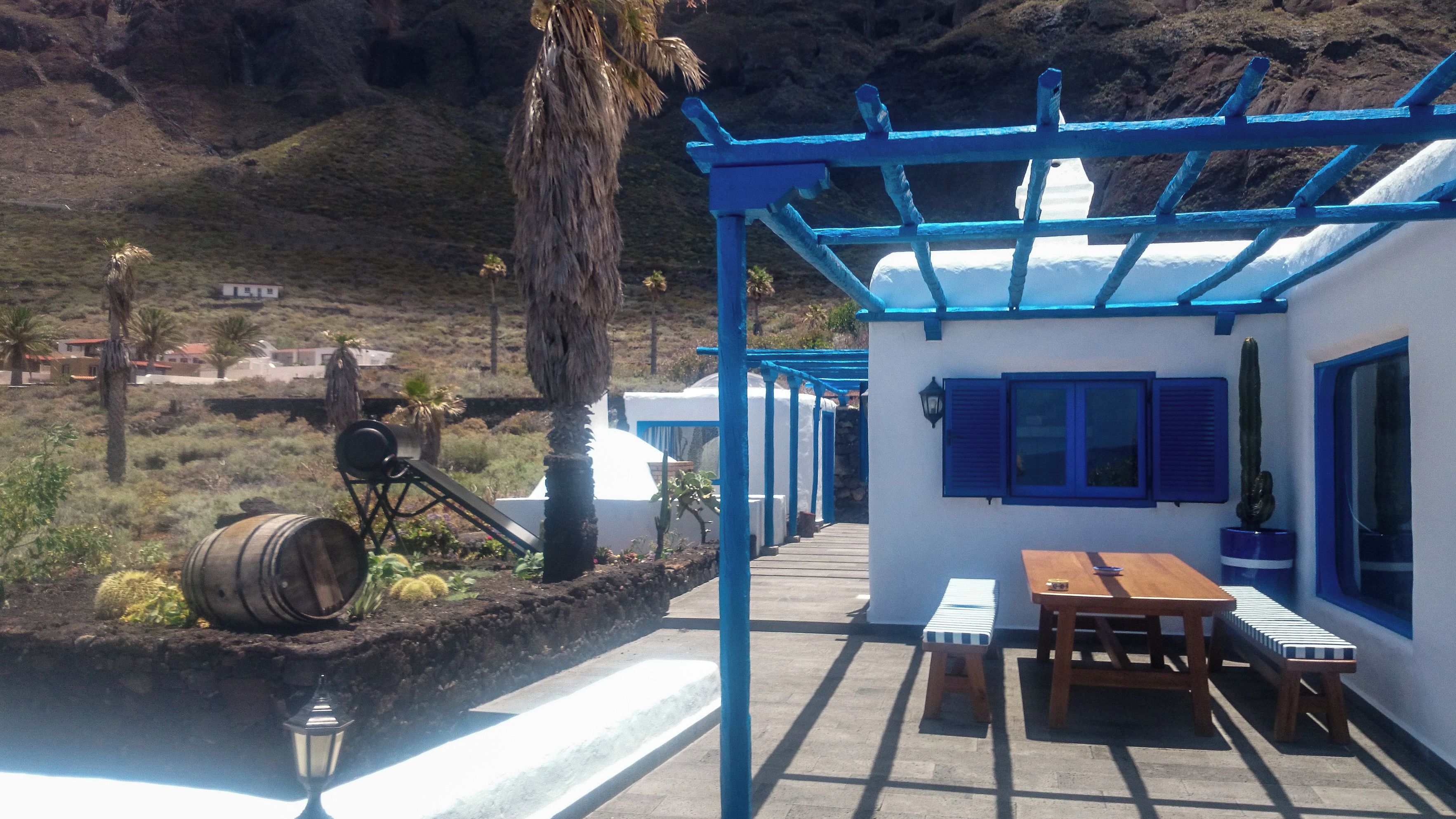 Un espacio de estar al aire libre cubierto con una mesa y bancos de madera, una pérgola azul y vistas a un paisaje montañoso.