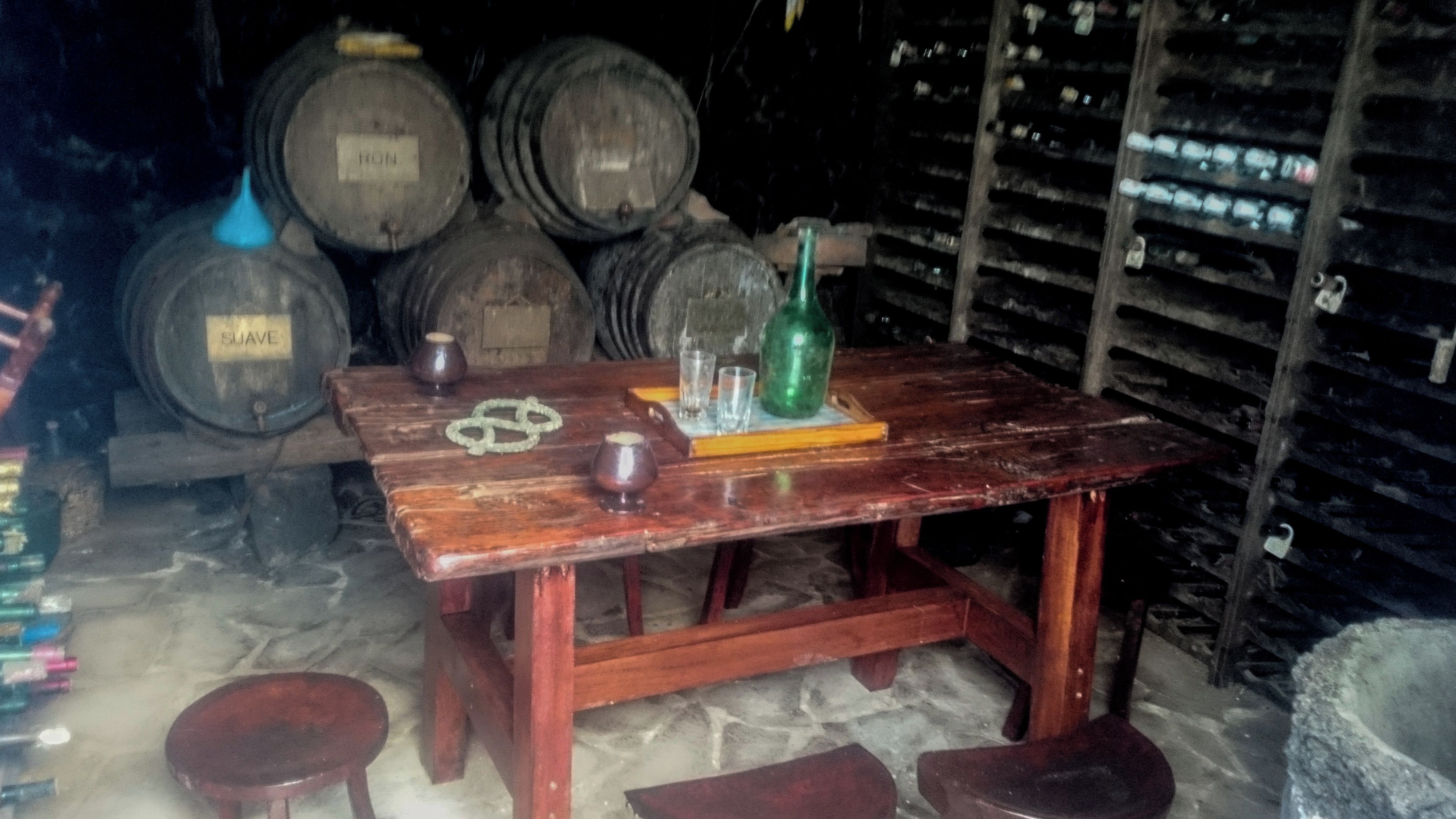 Una mesa de madera rústica con una botella y copas, rodeada de barriles en una bodega tenuemente iluminada.