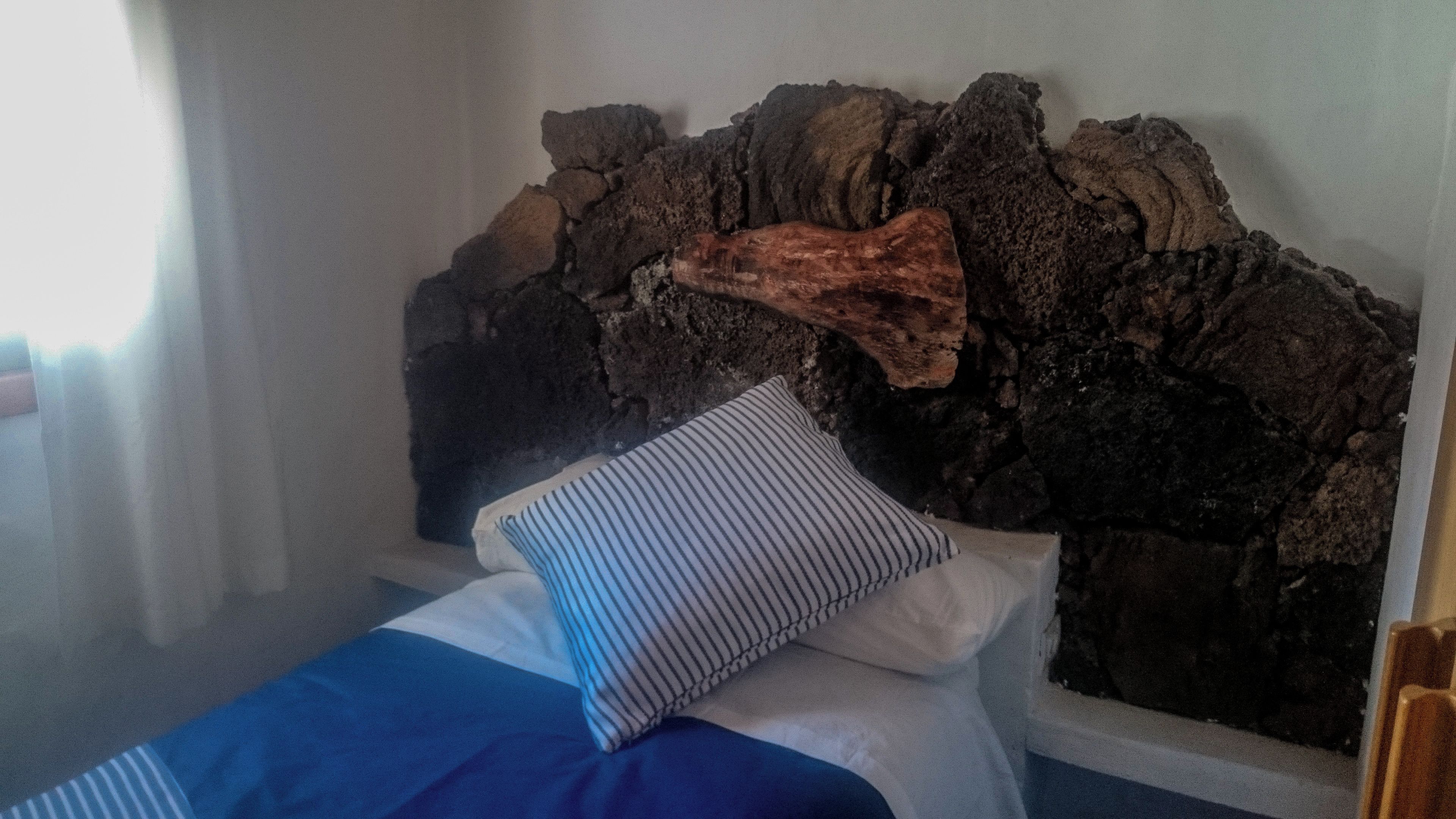 Una cama con un cojín a rayas azul y blanco y otro cojín blanco, frente a una pared con una superficie rocosa y rugosa.