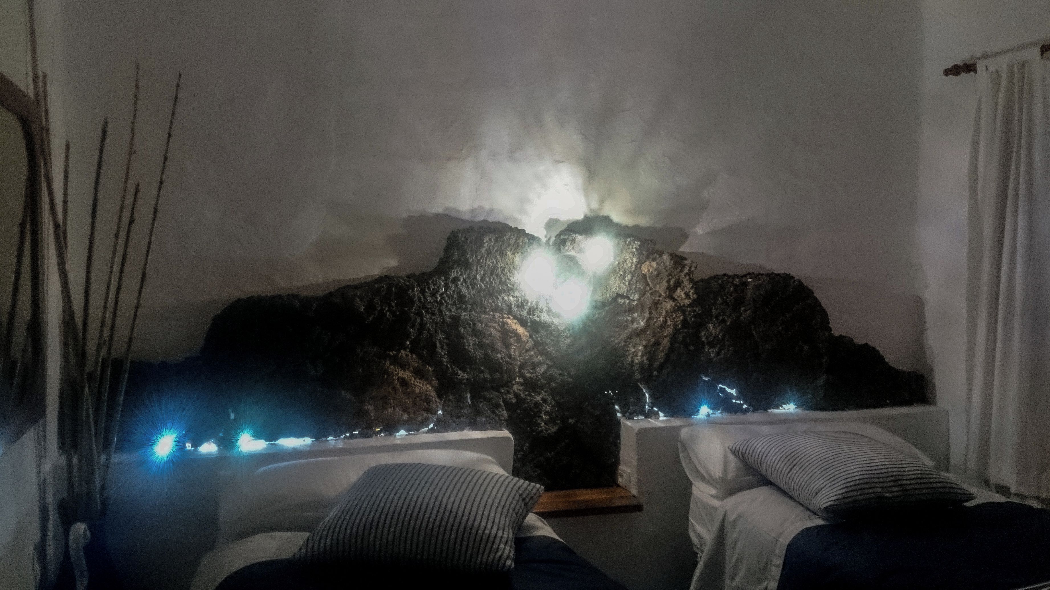 A room with two beds, a rock formation with lights, and a view of the sea.