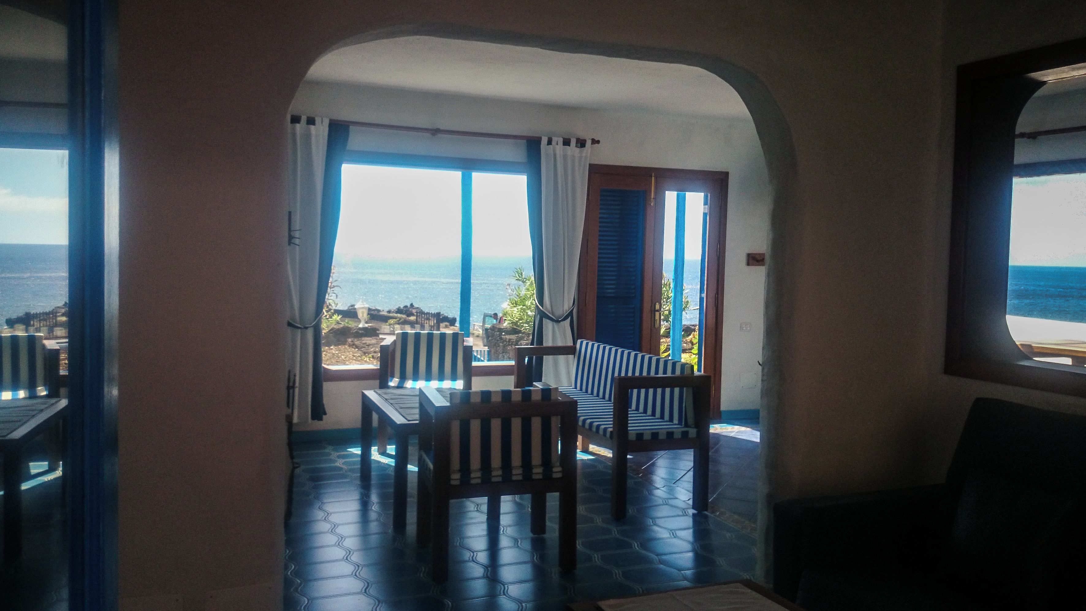 Una habitación con vista al mar, que incluye una mesa, sillas, una ventana con cortinas y piso de baldosas.