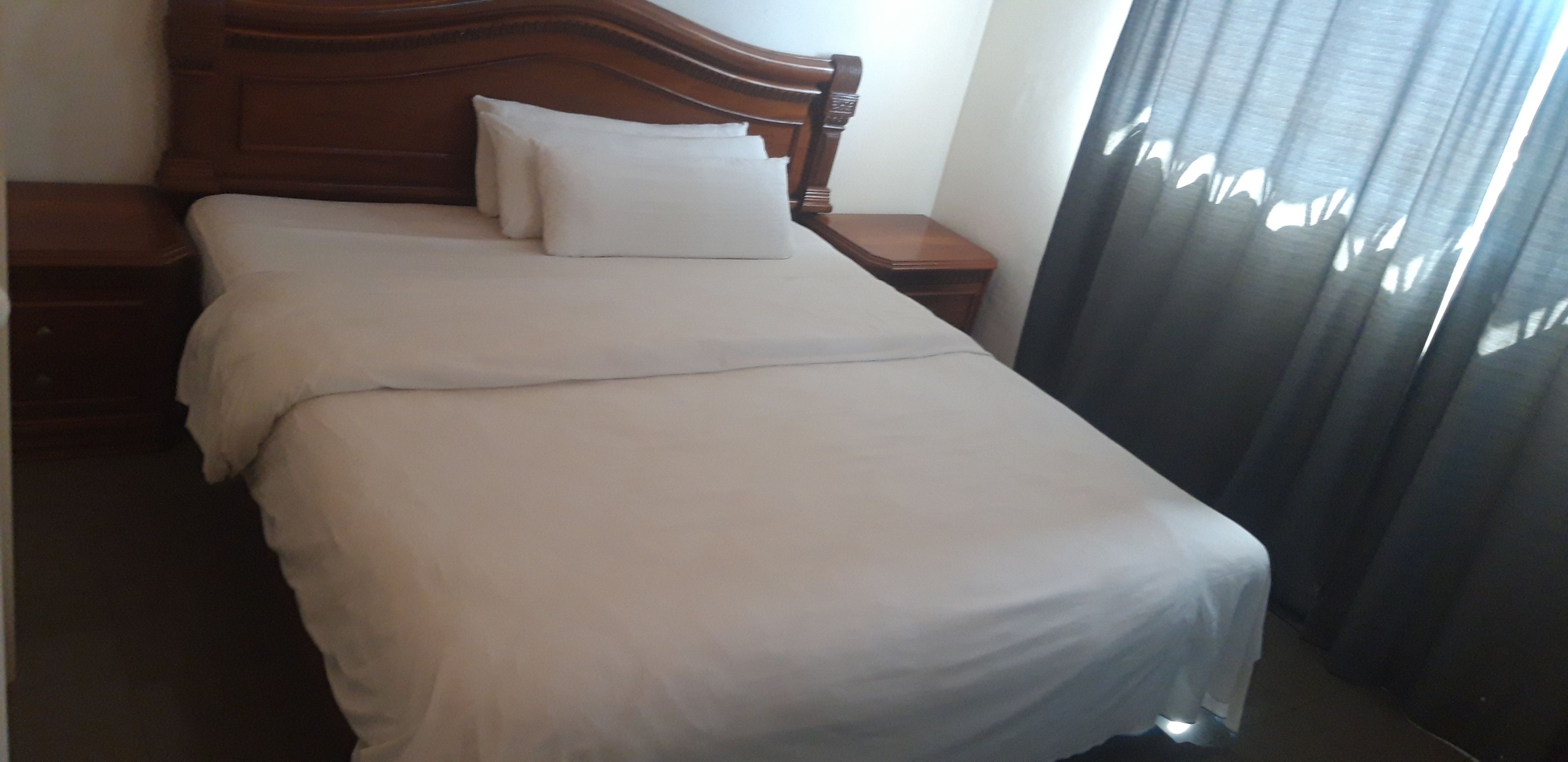 A neatly made bed with white linens and pillows, a wooden headboard, and a nightstand with a lamp.
