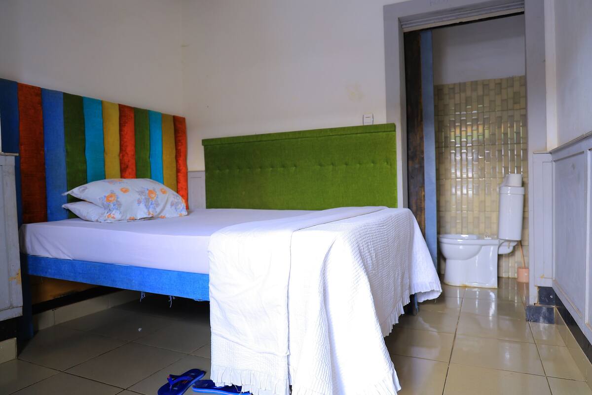 A room with a bed, a green headboard, a white bedspread, a white blanket, a white pillow, a blue pair of flip-flops, a white toilet, and a tiled floor.