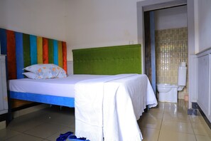 A room with a bed, a green headboard, a white bedspread, a white blanket, a white pillow, a blue pair of flip-flops, a white toilet, and a tiled floor. - Sweet Stay Guest House (Kigali)
