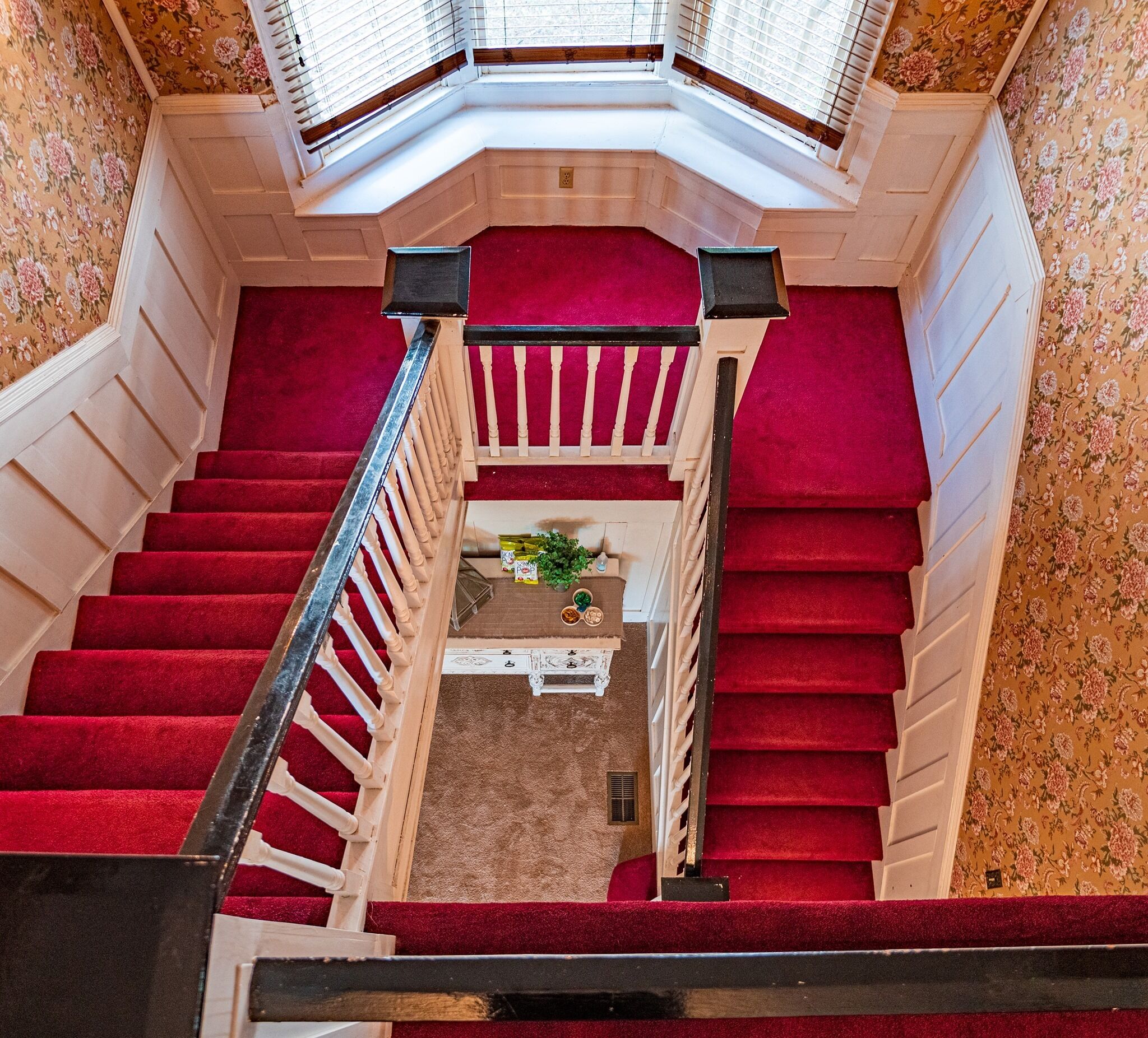A grand staircase with red carpeted steps and a wooden railing, leading to a landing with a small table and potted plant.