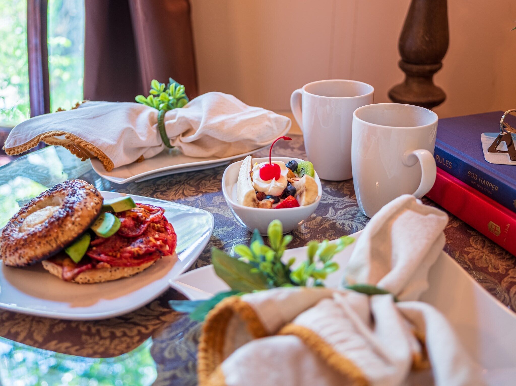 Mesa de café da manhã com uma bandeja de bagels, uma tigela de frutas e um livro chamado 'Rules' de Bob Harper.