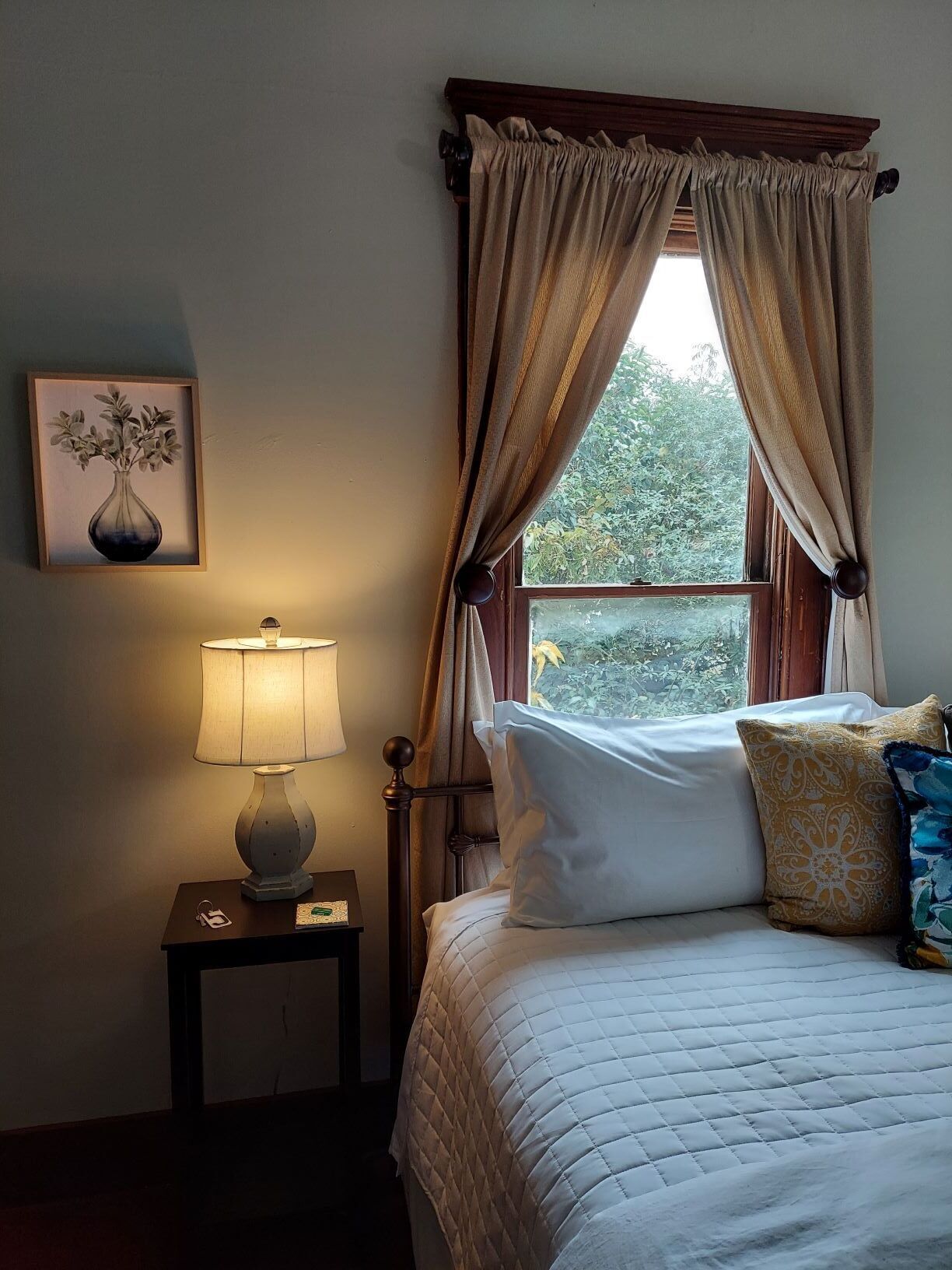 A bedroom with a bed, a nightstand with a lamp, a window with curtains, and a framed picture on the wall.