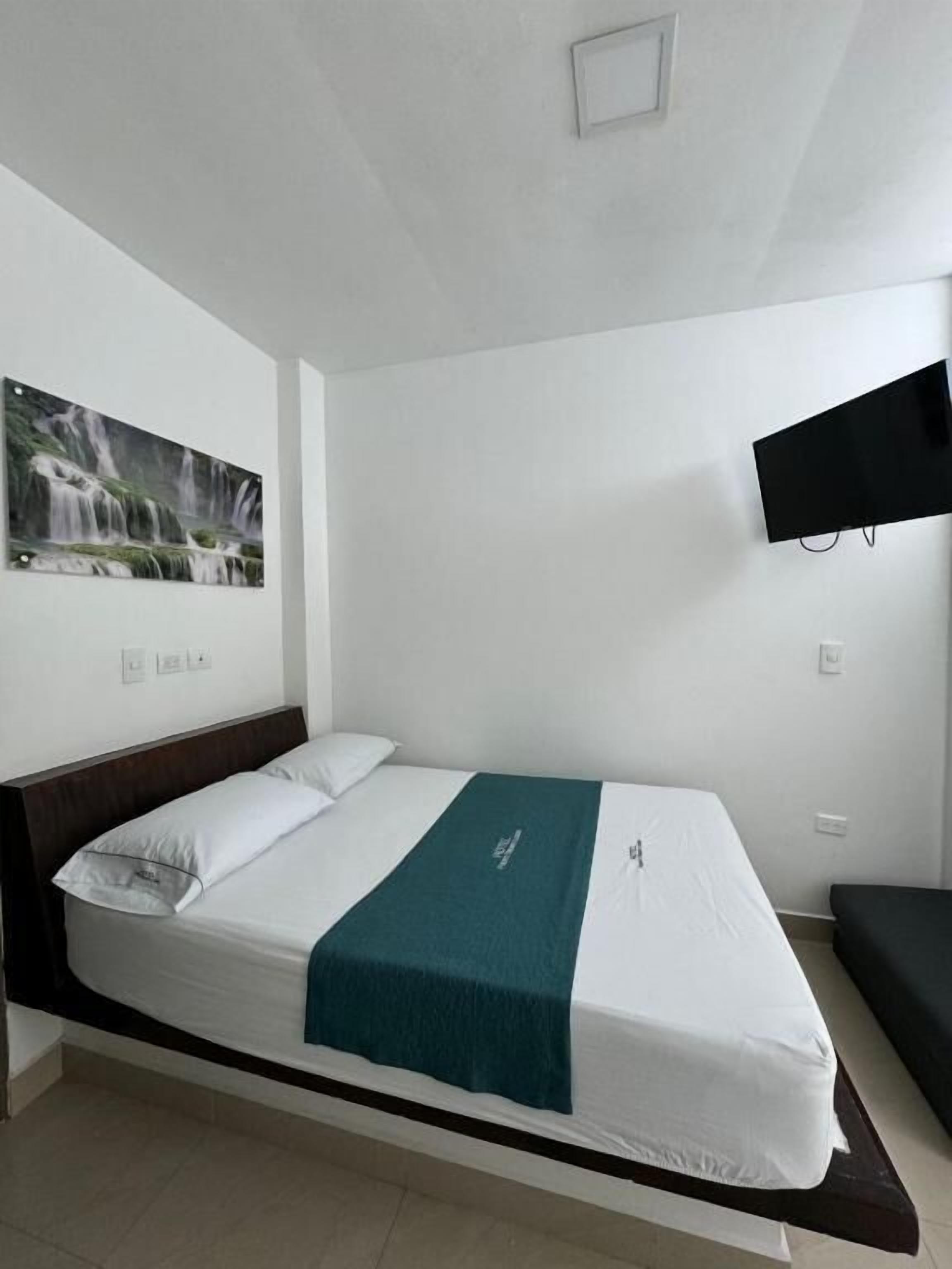 Una cama individual con colchón blanco y manta color turquesa, cabecero oscuro, televisor montado en la pared y un cuadro con una cascada.