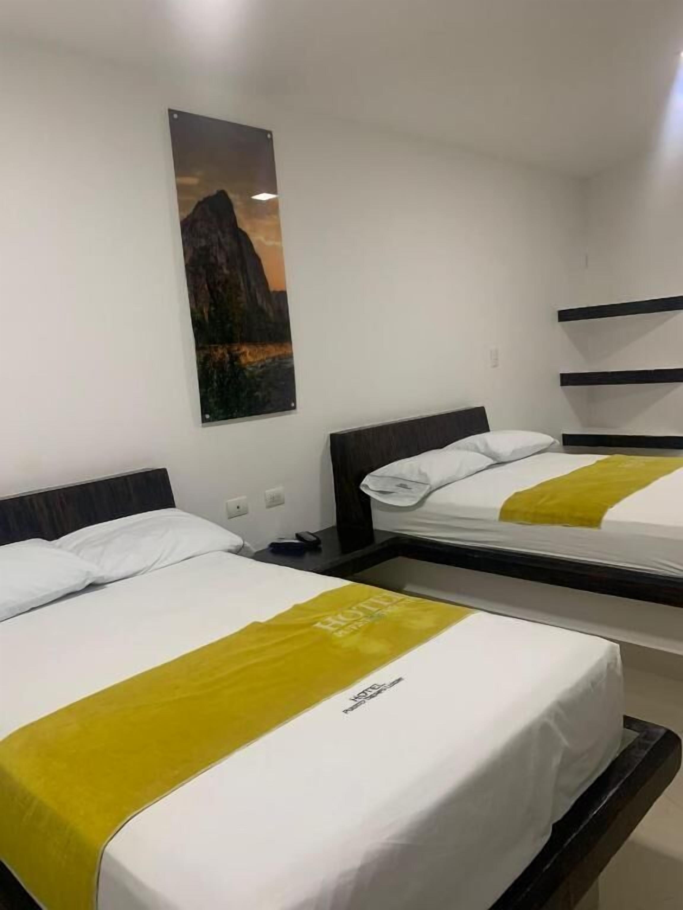 Une chambre d’hôtel avec deux lits, chacun agrémenté de draps blancs et d’un élément décoratif jaune. Un tableau encadré est accroché au mur, au-dessus des lits.