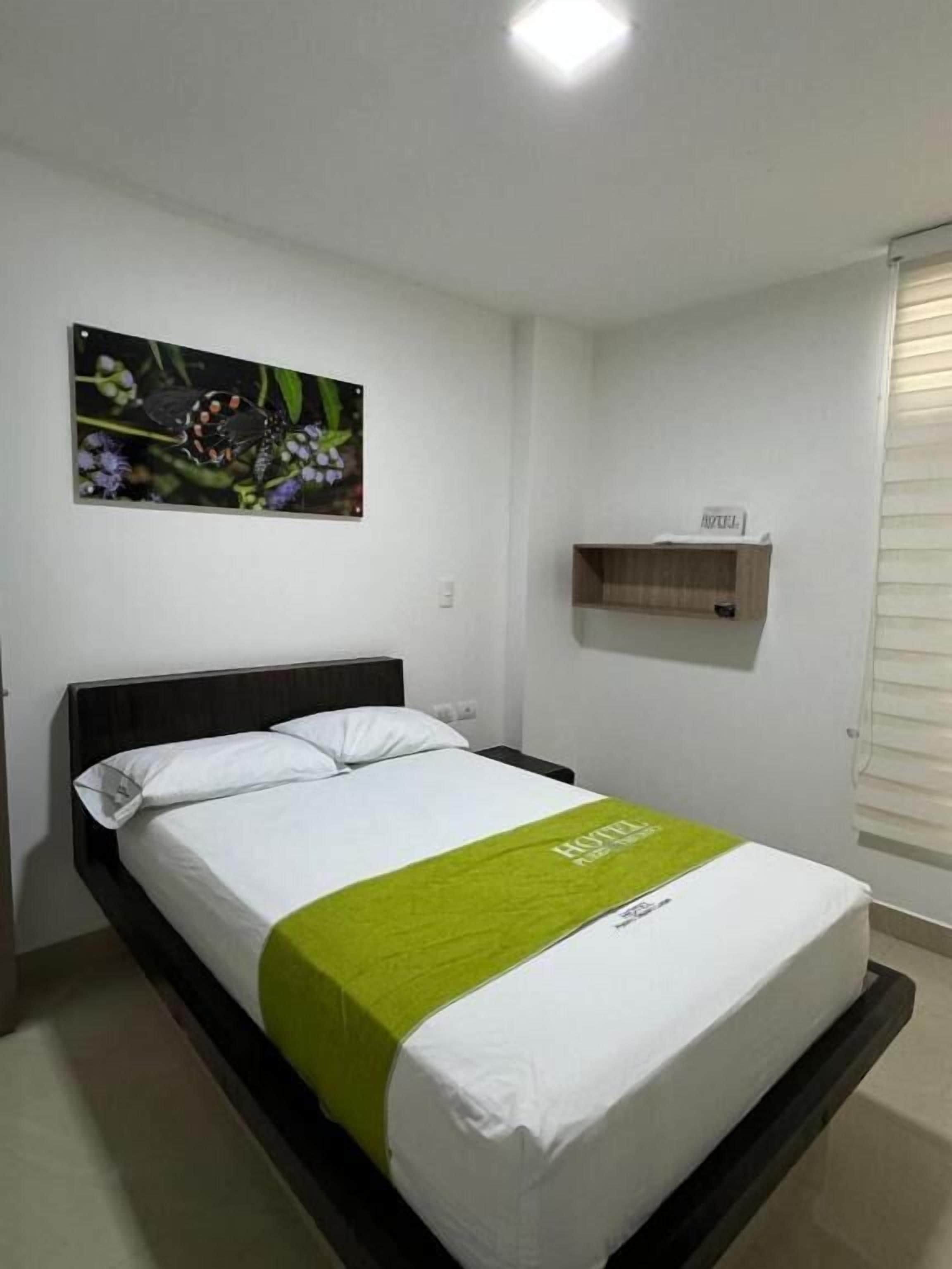 A hotel room with a bed, a green bedspread, a painting of flowers on the wall, a small wooden shelf, and a window with blinds.