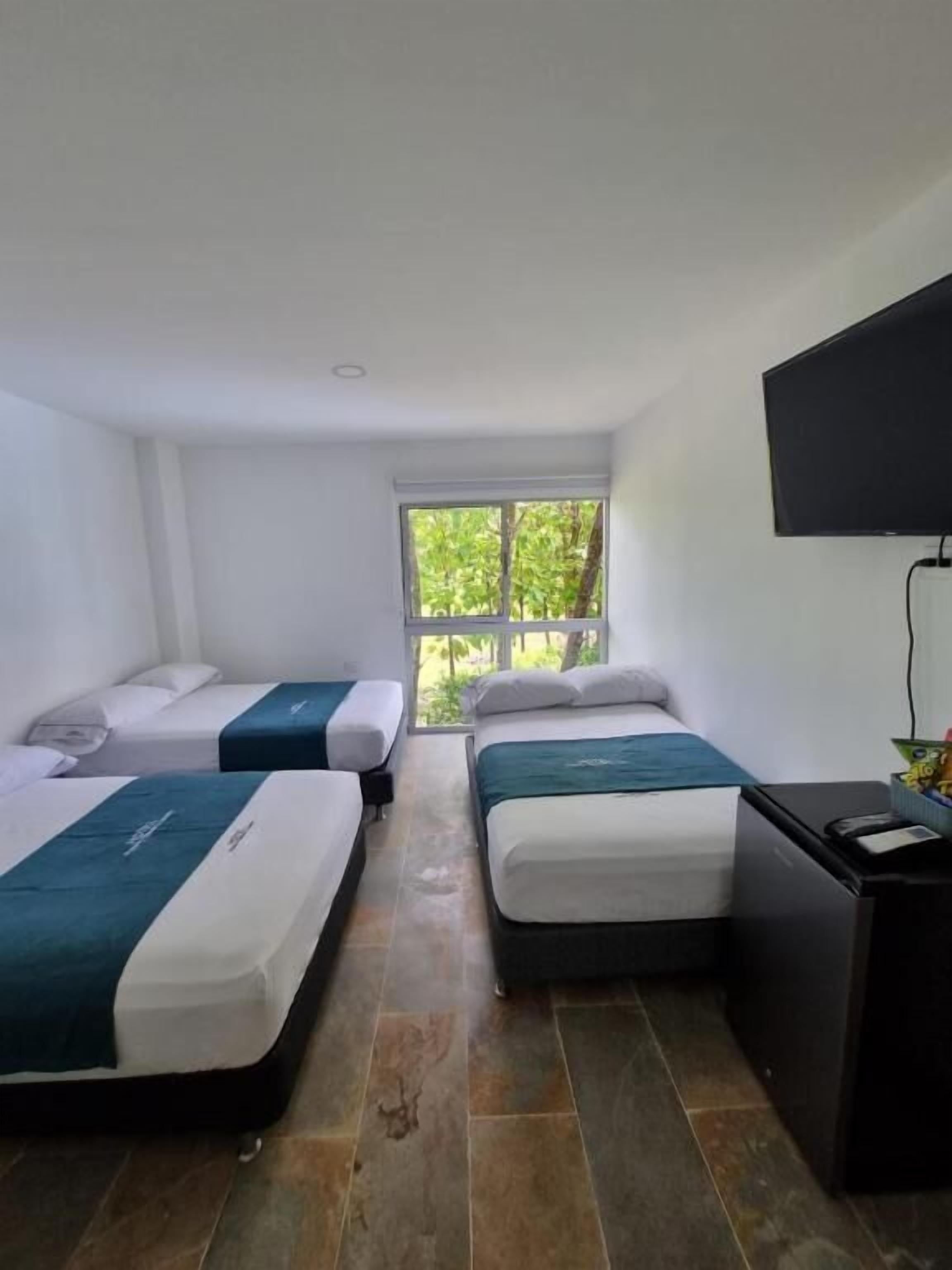Ein Hotelzimmer mit zwei Einzelbetten, einem kleinen Schreibtisch, einem Fernseher und einem Fenster mit Blick auf Bäume.