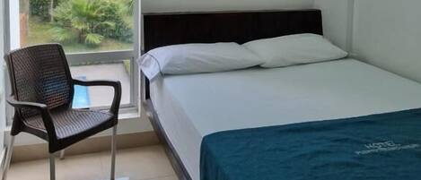 A hotel room with a bed, a chair, a window with a view of greenery, and a painting on the wall.