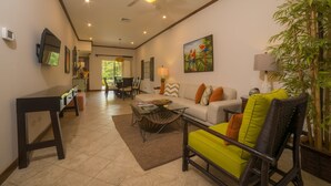 A spacious living room with a sofa, armchairs, a coffee table, and a television. There is a ceiling fan, a painting, and a lamp on a side table. - Los Suenos Resort Veranda 8F (Jacó)