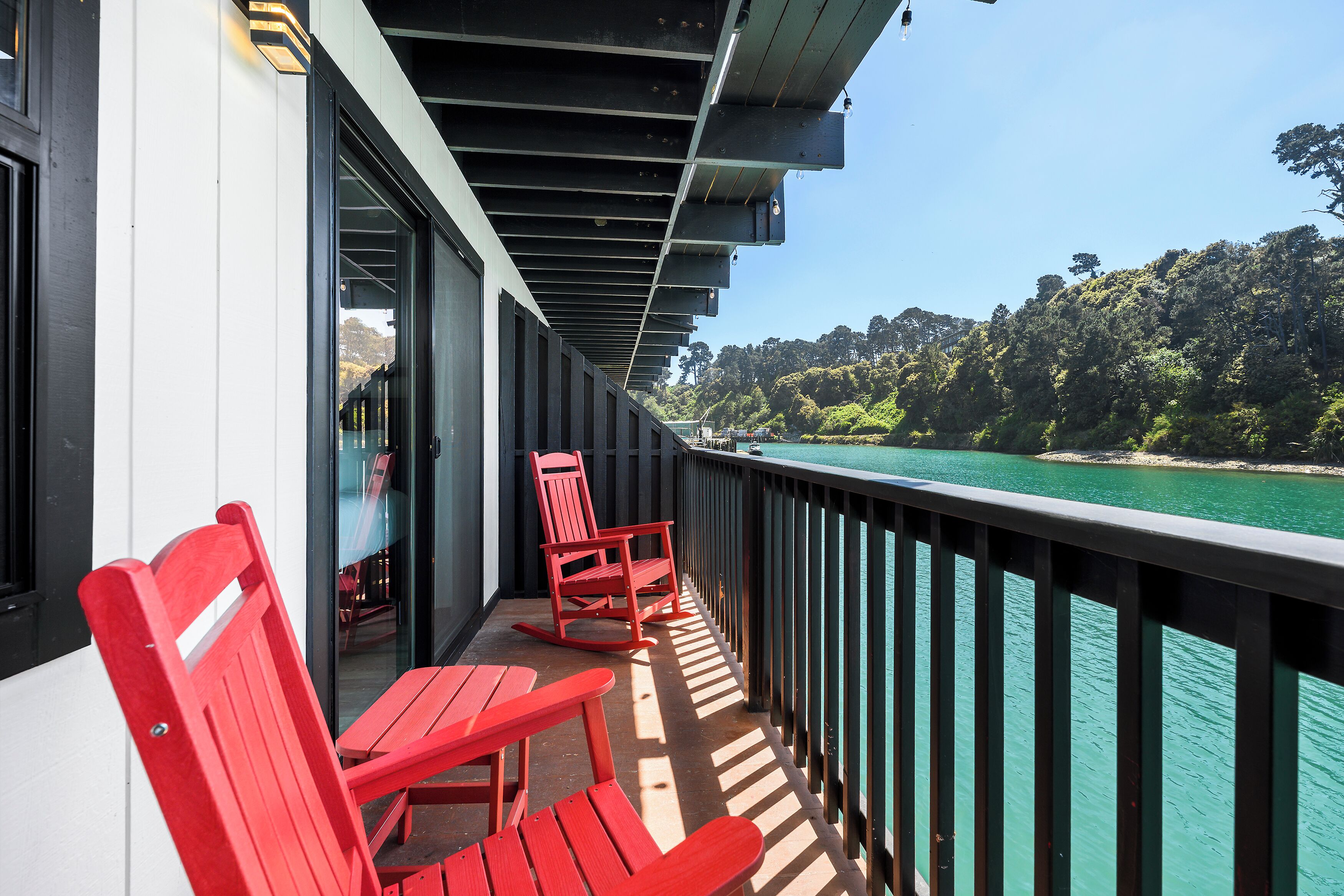 A balcony with red chairs, a table, and a view of a forested coastline.