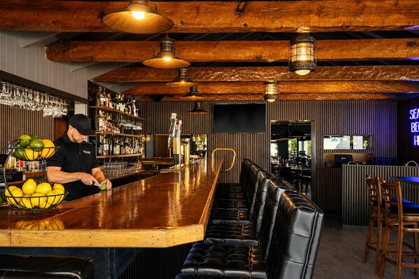 A bar area with a bartender, a bar counter, stools, and a shelf with bottles and glasses. - Anchor Lodge (Fort Bragg)