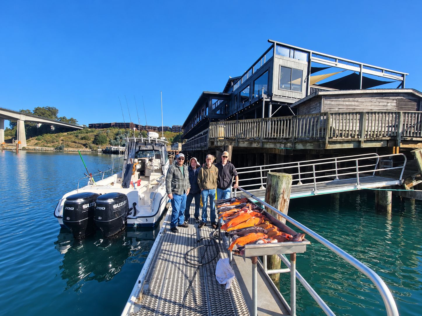 A boat docked at a pier with a wooden structure and a bridge in the background.