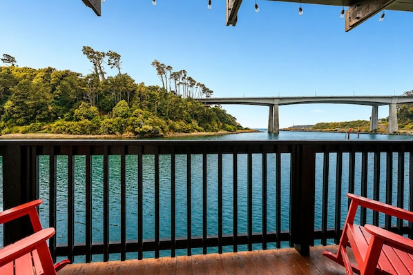 A wooden deck with a railing overlooking a body of water, a bridge in the distance, and a forested island. - Anchor Lodge (Fort Bragg)