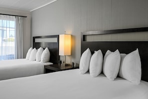 A hotel room with two beds, a nightstand with a lamp, and a view of the outdoors. - Anchor Lodge (Fort Bragg)