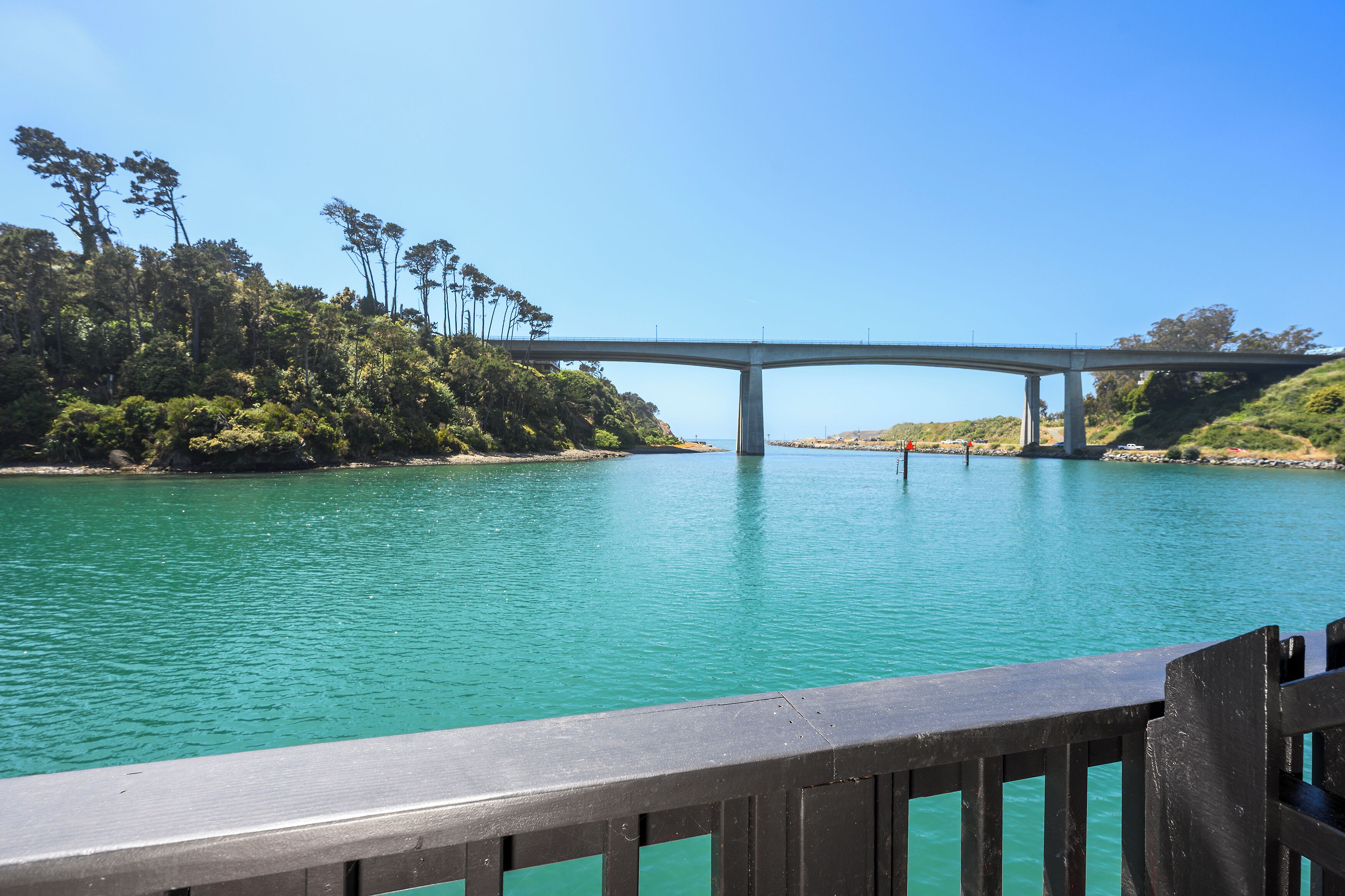 Een brug over een turquoise waterpartij, met een beboste helling aan de ene kant en een helderblauwe lucht erboven.