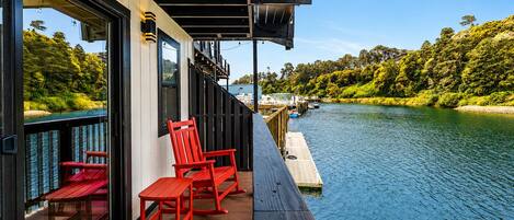 A deck with red chairs and a table overlooking a river, with a dock extending into the water.