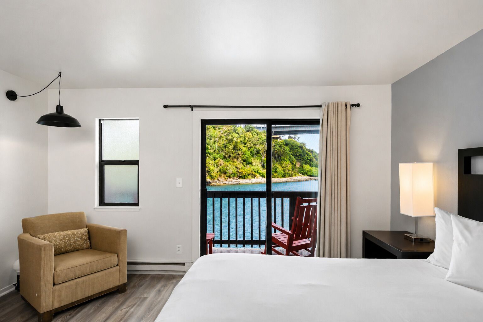 A hotel room with a bed, a chair, a nightstand, a lamp, a window, and a view of a balcony with a railing.