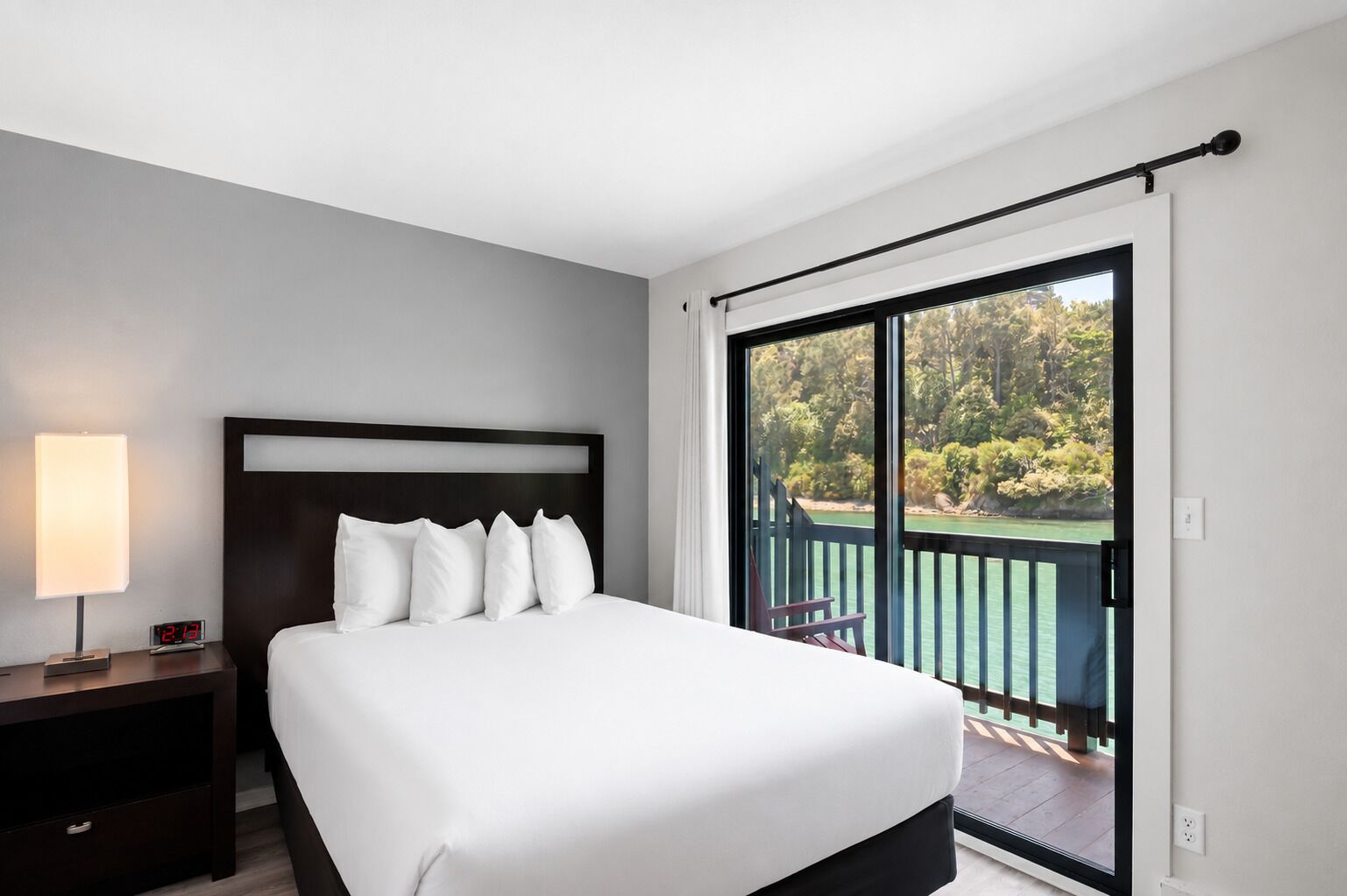 Une chambre d’hôtel avec un lit, une table de chevet, une lampe et une vue sur un balcon doté d’une balustrade.