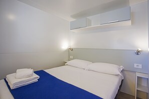 A hotel room with a bed, white linens, a blue bedspread, a white towel stack, a nightstand, a wall-mounted TV, and a wall-mounted light. - Playa Colorada (Gaeta)