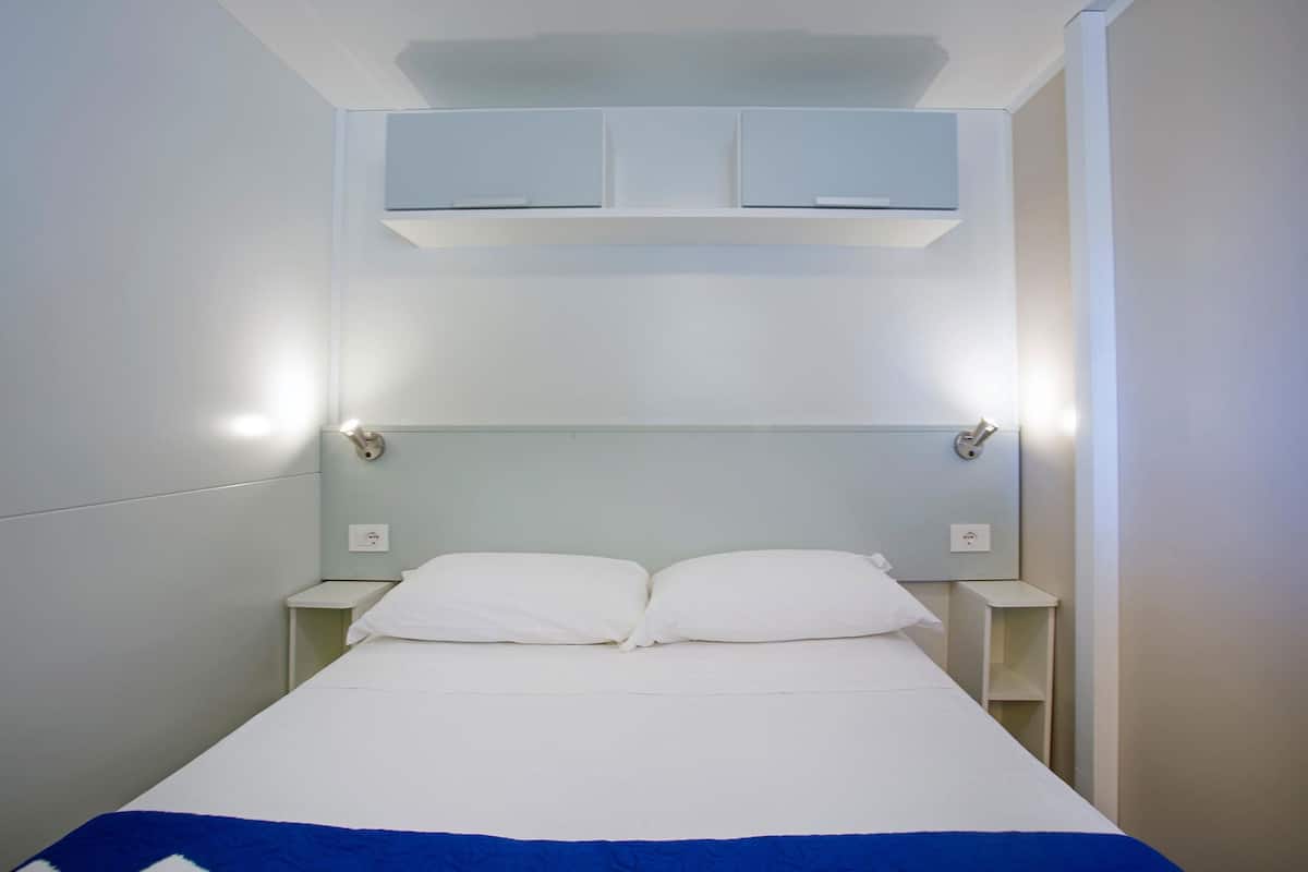 Uma cama de solteiro com roupa de cama branca e um detalhe azul no rodapé. Existem duas mesas de cabeceira com abajures, e uma prateleira fixada na parede acima da cama.