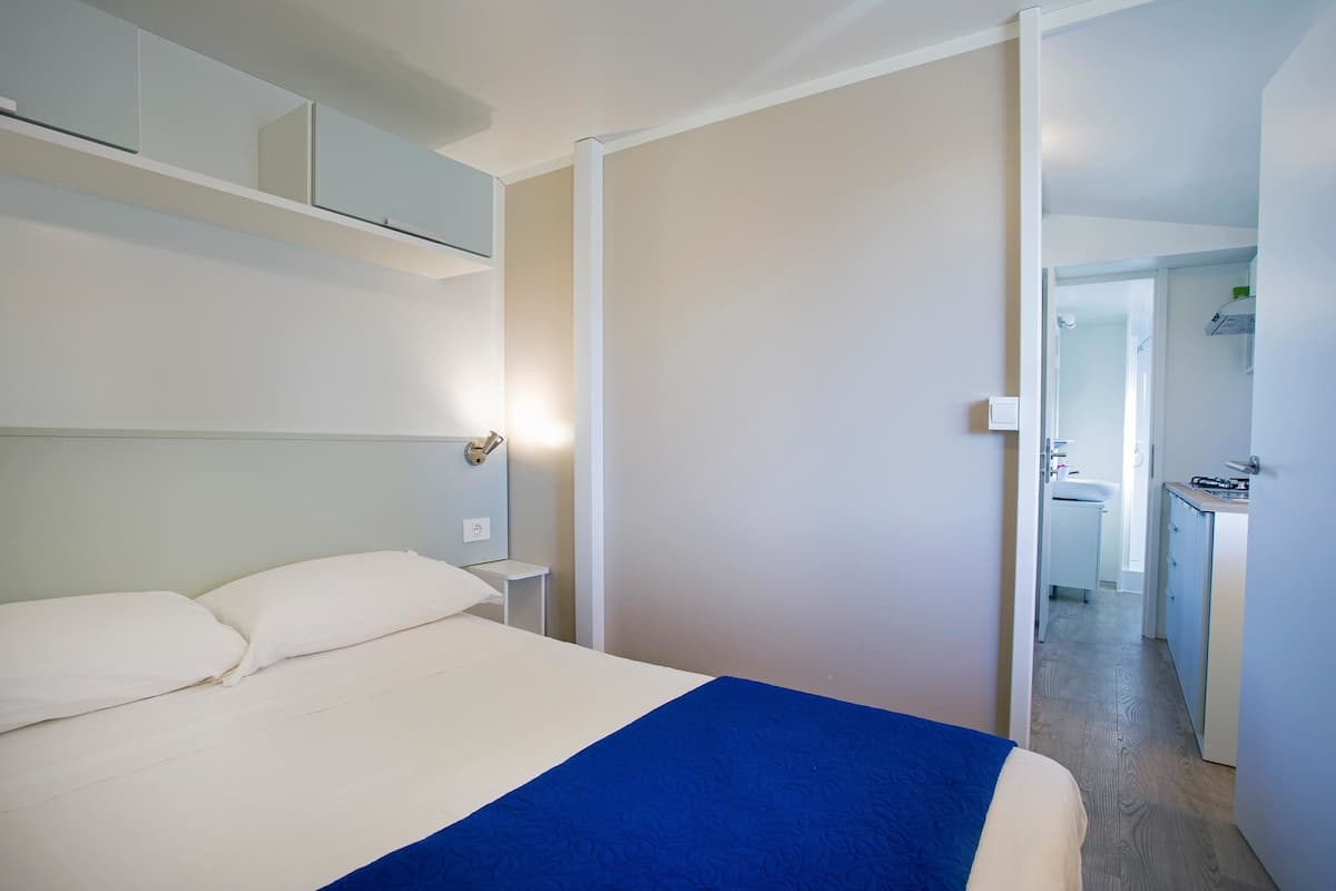 Quarto de hotel com cama, mesa de cabeceira, espelho e vista para o banheiro e cozinha.