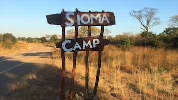 Um sinal rústico com a inscrição "SIOMA CAMP", em um caminho de terra ao lado de uma estrada, cercado por grama seca e árvores esparsas.