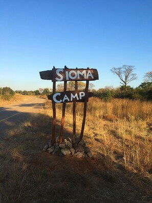 A rustic signpost with "SIOMA CAMP" written on it, standing on a dirt path beside a road, surrounded by dry grass and sparse trees. - Sioma Camp (Sioma)