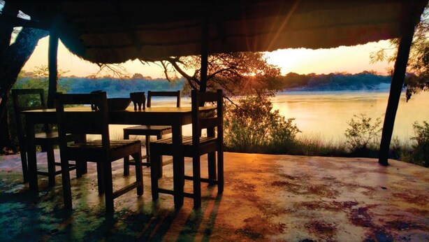 An outdoor dining area with a table and chairs, overlooking a calm body of water and a distant landscape during sunset.