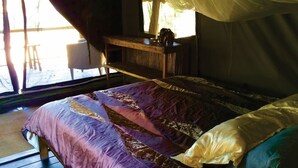 A bed with a purple and gold quilt, a wooden nightstand with a kettle, and a window with a white curtain.