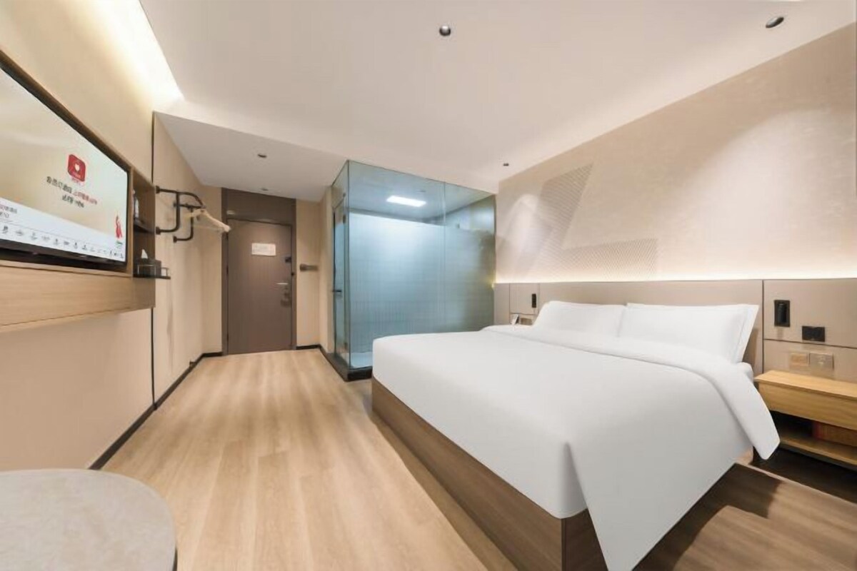 A modern hotel room with a large bed, a TV on the wall, and a glass partition separating the room from the hallway.
