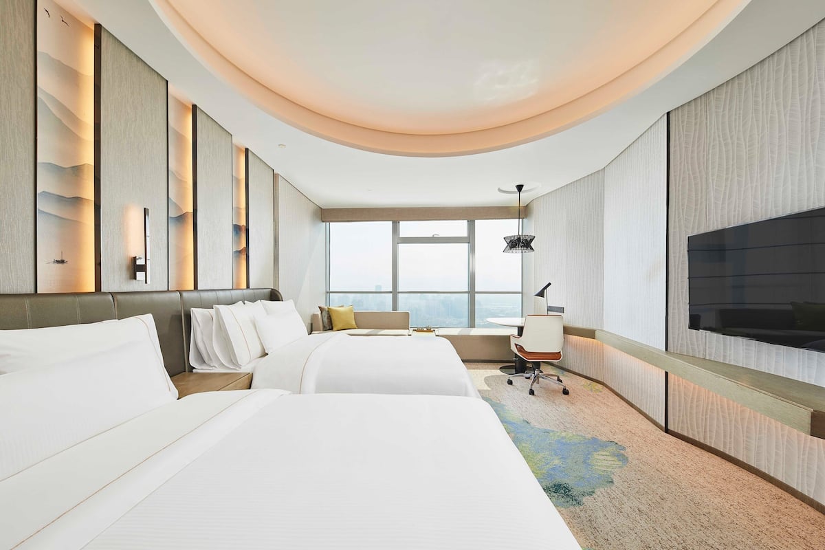 Ein Hotelzimmer mit zwei Betten, einem Schreibtisch, einem Sessel, einem Fernseher und einem großen Fenster mit Blick auf die Stadt.