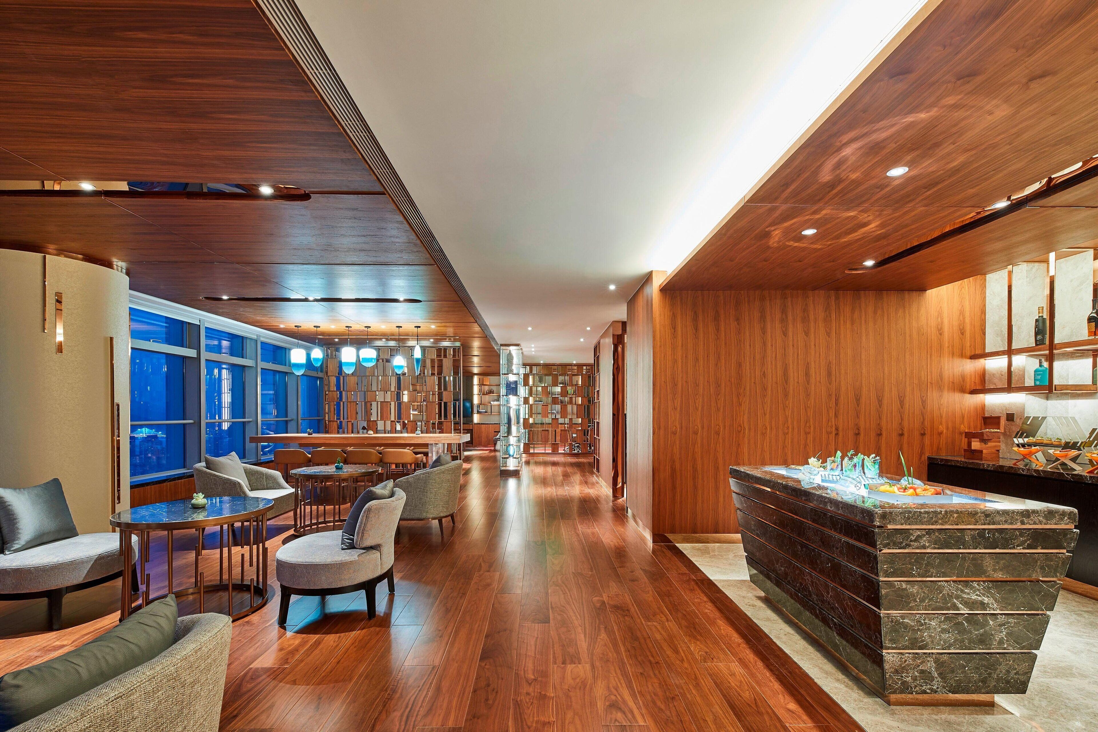 Een moderne hotellobby met houten vloeren, zithoeken, een receptiebalie en uitzicht op de stad door grote ramen.
