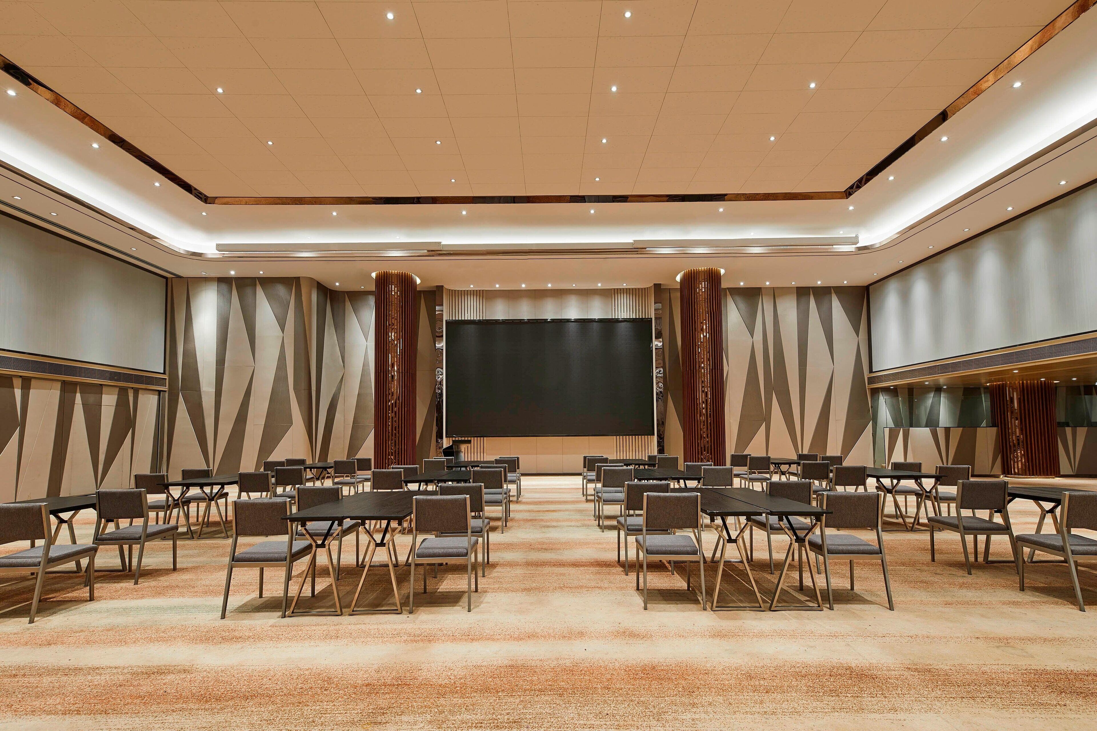 Un salón de conferencias con filas de sillas y mesas, una pantalla grande al frente y un diseño de pared con motivos.