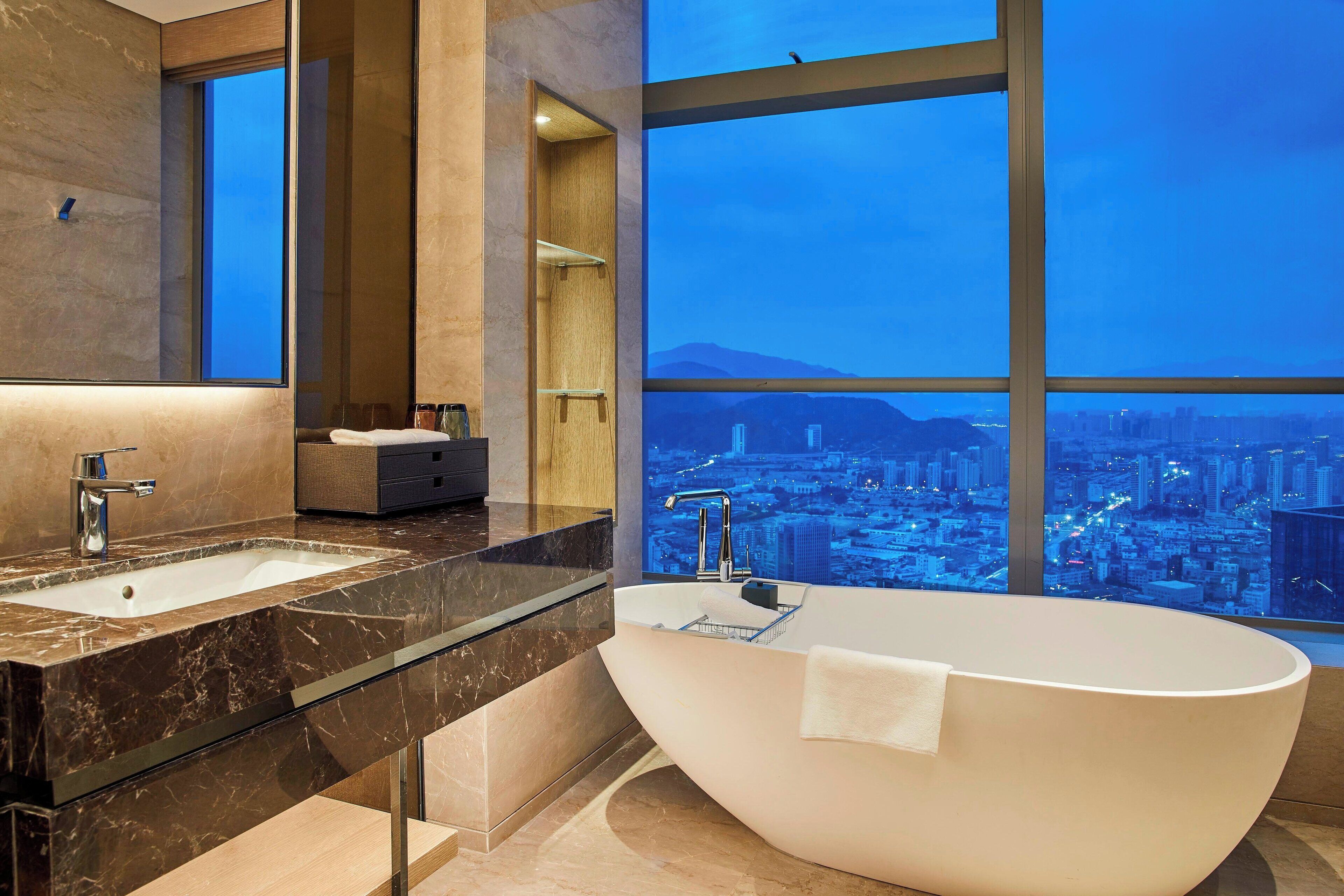 A modern bathroom with a large bathtub, two sinks, and a city view through large windows.