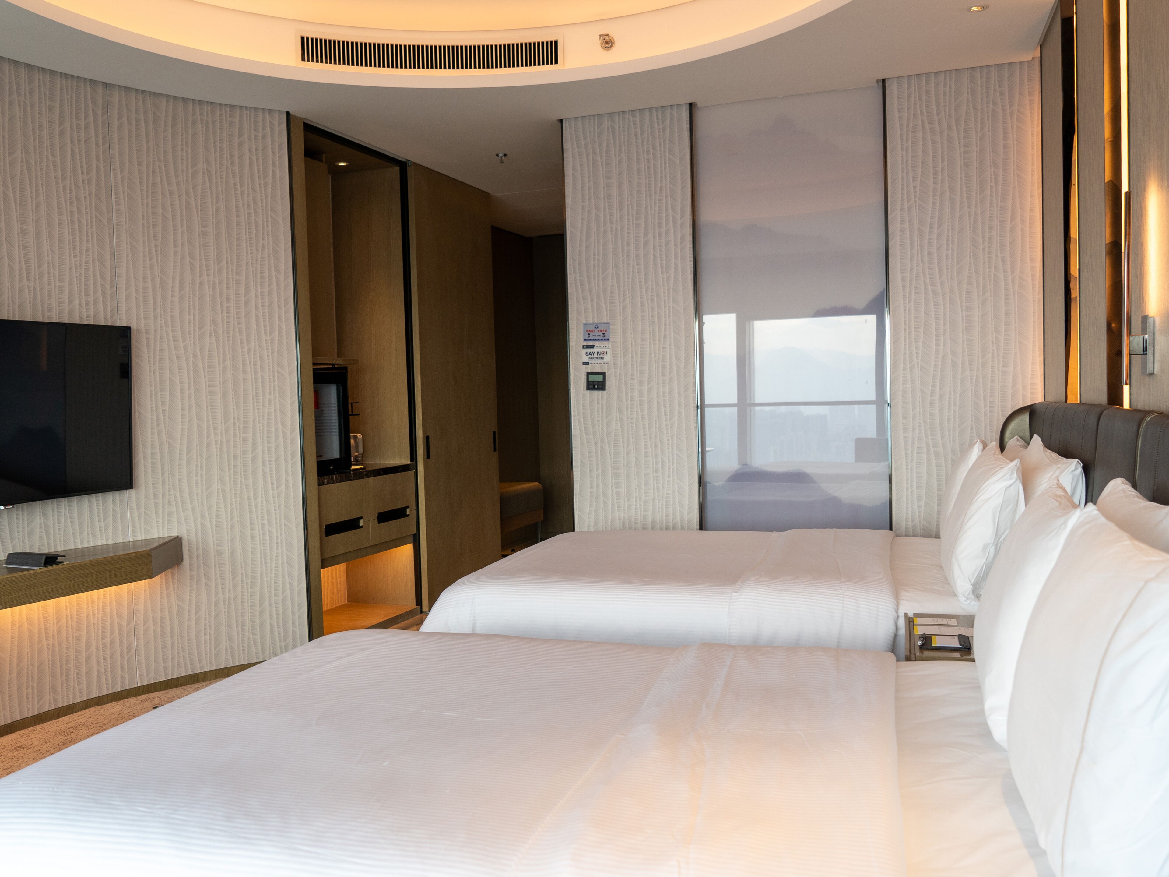 A hotel room with two beds, a TV, a desk, and a view of the city.
