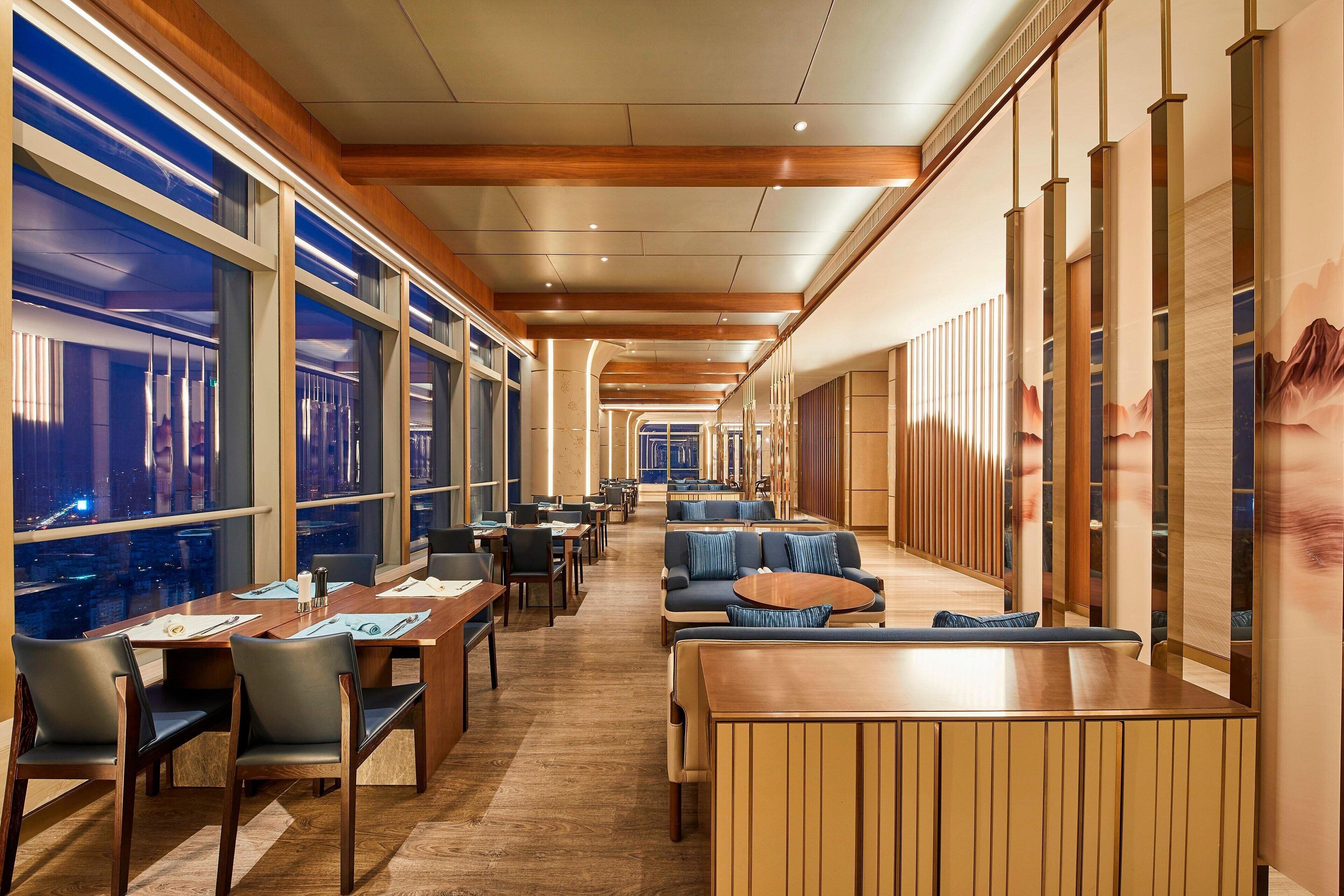 A modern hotel lobby with wooden flooring, seating areas, and large windows offering city views.